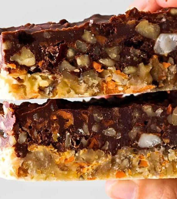 Hand holding two stacked slices of a chocolate and nut dessert bar, with visible layers of nuts, chocolate, and other ingredients.