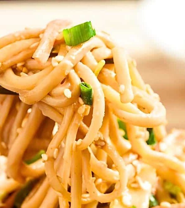 Chopsticks holding up a portion of stir-fried noodles with green onions and sesame seeds, with a dish of sauce in the background.