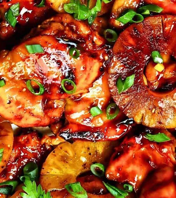 Close-up of a dish featuring glazed grilled chicken pieces topped with pineapple slices, chopped green onions, and parsley leaves.