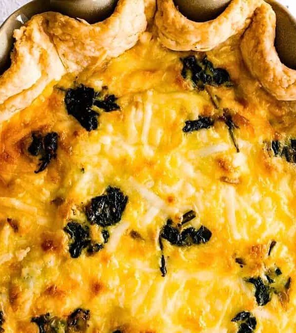 A baked quiche with flaky crust, filled with a creamy mixture containing cheese and spinach, displayed in a pie dish.