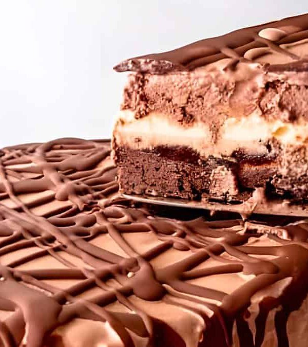 A chocolate, vanilla, and caramel layered ice cream cake with chocolate drizzles. One slice is being lifted from the whole cake.