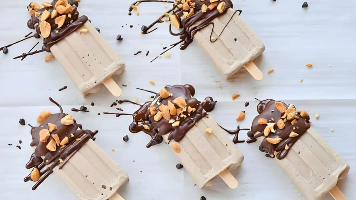 Five popsicles are arranged on a white background, each dipped in chocolate and topped with chopped nuts, with additional chocolate drizzle and scattered nuts.
