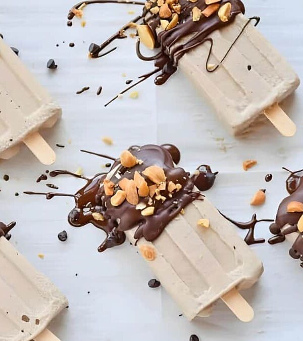 Five popsicles are arranged on a white background, each dipped in chocolate and topped with chopped nuts, with additional chocolate drizzle and scattered nuts.