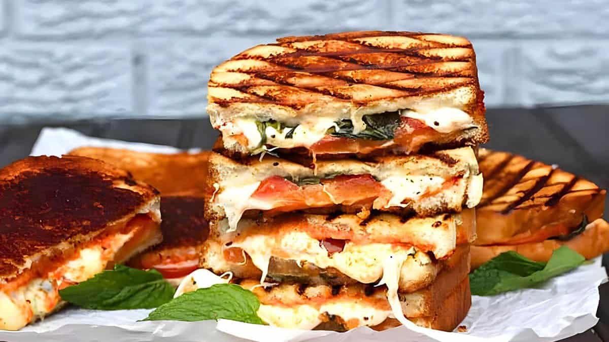 A stack of grilled cheese sandwiches filled with tomatoes, spinach, and melted cheese, set on a white parchment paper background with a brick wall backdrop. Fresh mint leaves are scattered around.