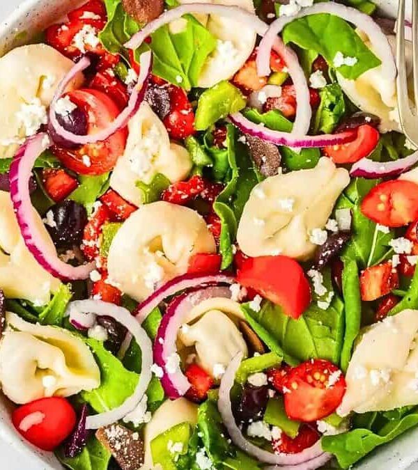 A bowl of fresh salad featuring tortellini, spinach, cherry tomatoes, black olives, and sliced red onions, garnished with crumbled feta cheese. A fork rests on the edge of the bowl.