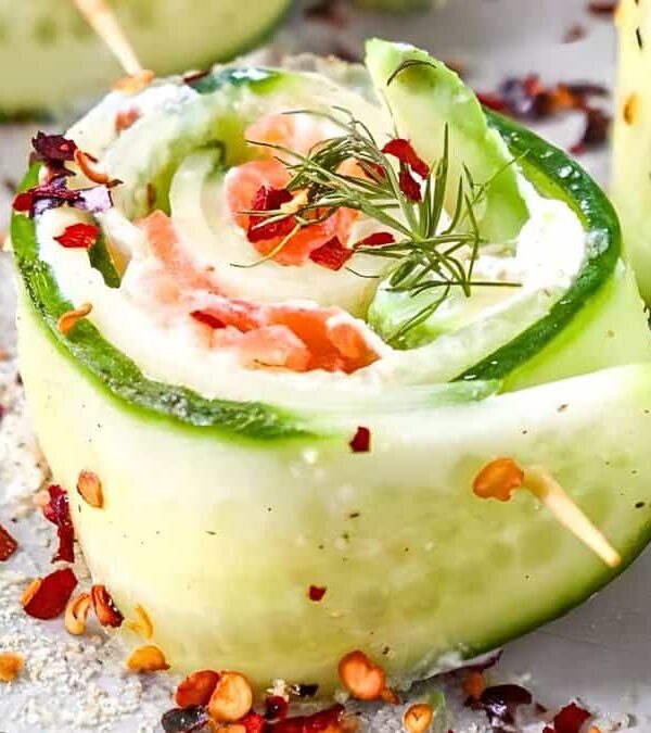 Close-up of a cucumber roll filled with salmon, cream cheese, and garnished with dill and red pepper flakes. Toothpick holds the roll together, surrounded by scattered spices.