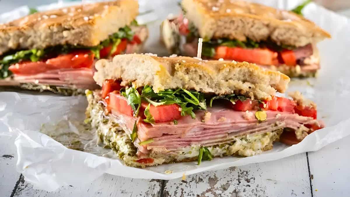 A close-up of a cut sandwich with layers of ham, tomato, lettuce, and spread on a sesame-seed bun, placed on parchment paper.