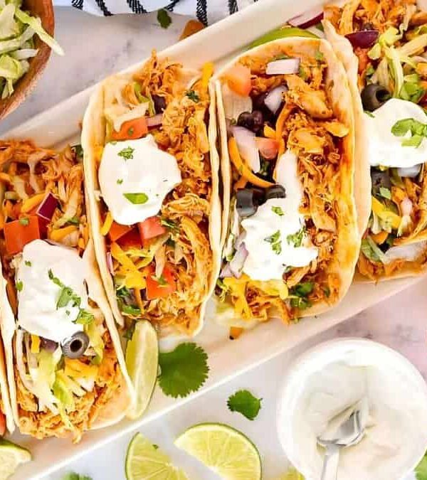 A platter of four tacos filled with shredded meat, vegetables, cheese, olives, and sour cream. Lime wedges and a bowl of shredded lettuce are on the side. Fresh cilantro and tomatoes are in the foreground.