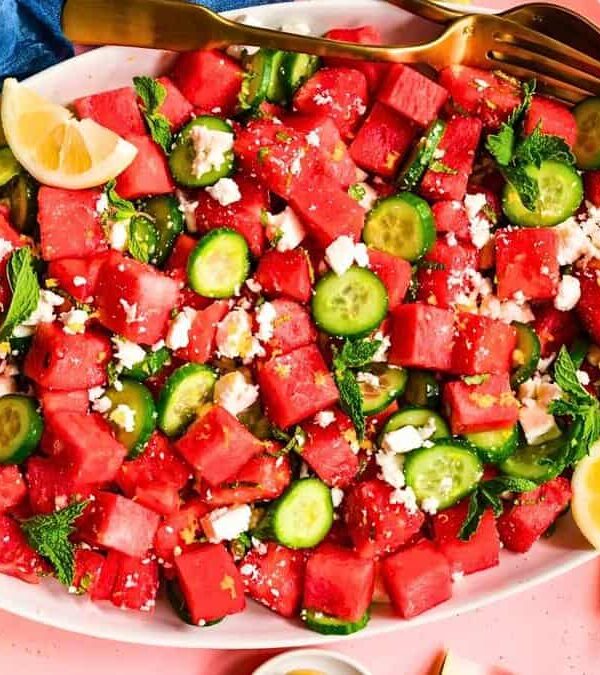 A platter of watermelon feta salad with cucumber slices, mint leaves, and lemon wedges on a pink surface. A fork and a spoon rest on top of the salad.