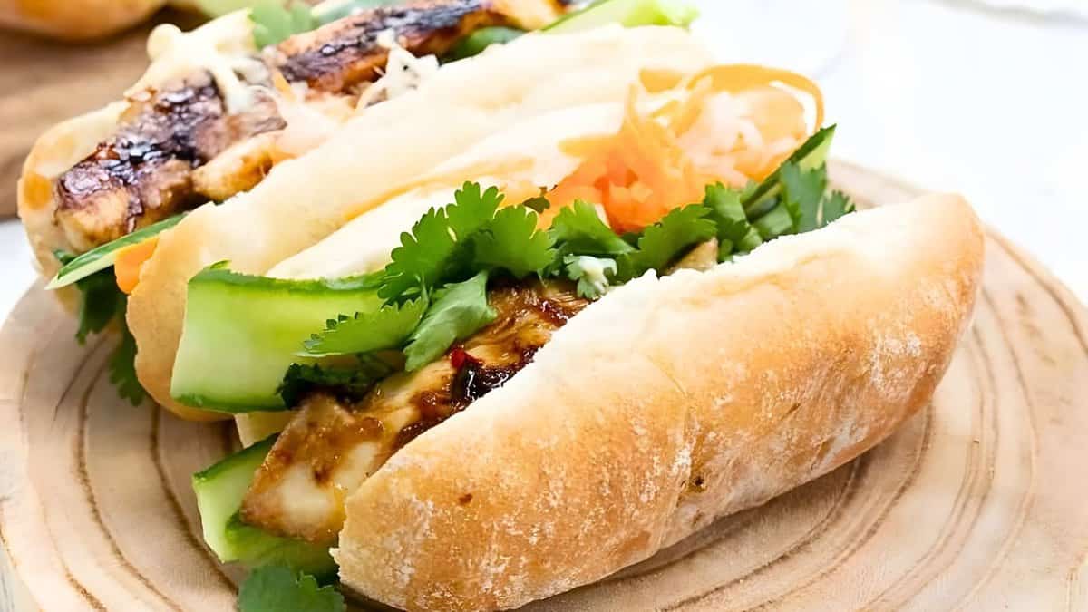 Two bánh mì sandwiches filled with grilled meat, fresh cilantro, cucumber slices, and pickled vegetables on a wooden plate.