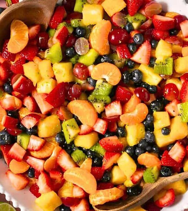 A bowl of colorful fruit salad containing strawberries, grapes, blueberries, pineapple, kiwi, and orange slices, with wooden serving utensils. Nearby, there are lime halves and a small bowl of zest.