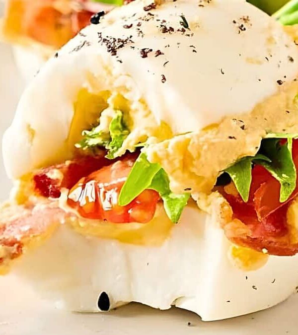 Close-up of a deviled egg topped with bacon, lettuce, tomato, and a sprinkle of black pepper, served on a white plate.
