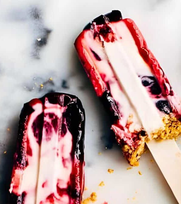 Four cherry cheesecake popsicles, some partially eaten, lay on a marble surface next to two fresh cherries.