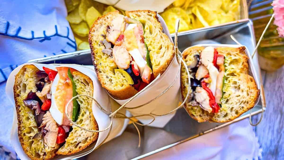 Three neatly wrapped sandwiches filled with meats, vegetables, and cheese, served on a tray with a side of potato chips.