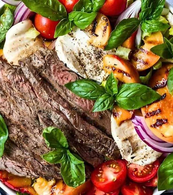 A bowl filled with slices of grilled steak, tomatoes, peaches, mozzarella, red onions, and fresh basil leaves.