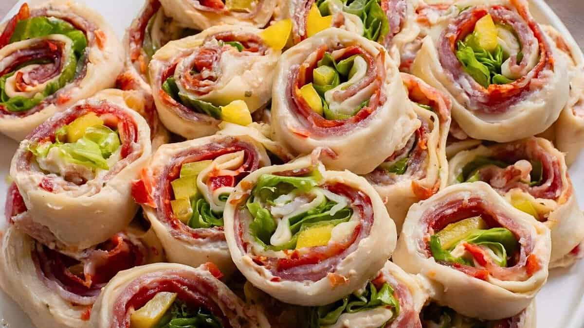 A plate filled with assorted pinwheel sandwiches made of rolled tortillas containing meats, cheese, lettuce, and yellow peppers.