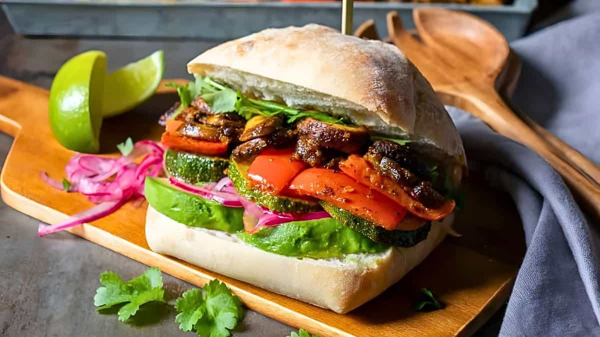 A ciabatta sandwich filled with avocado, red bell peppers, zucchini, seasoned mushrooms, and pickled onions, served on a wooden board with lime wedges and cilantro garnish. Wooden utensils are in the background.