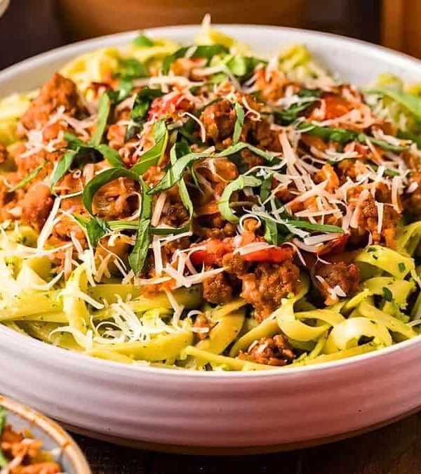 A bowl of pasta topped with meat sauce, grated cheese, and basil leaves, surrounded by additional dishes and ingredients on a table.