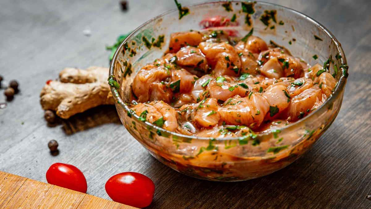 A glass bowl filled with marinated chicken pieces mixed with herbs and spices sits on a wooden surface. Ginger, cherry tomatoes, and peppercorns are scattered around the bowl.