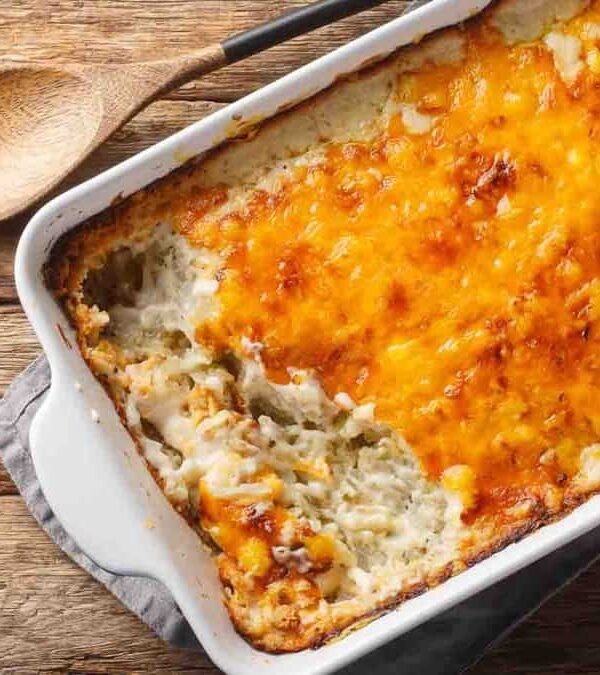 A rectangular white baking dish containing a partially served cheesy potato casserole, shown on a wooden surface with a wooden spoon beside it.
