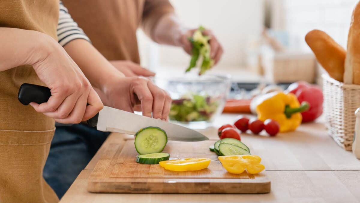 Two people cutting vegetables, including cucumbers and bell peppers, on a wooden cutting board in a kitchen. One person is holding a knife, and another is adding greens to a bowl in the background.