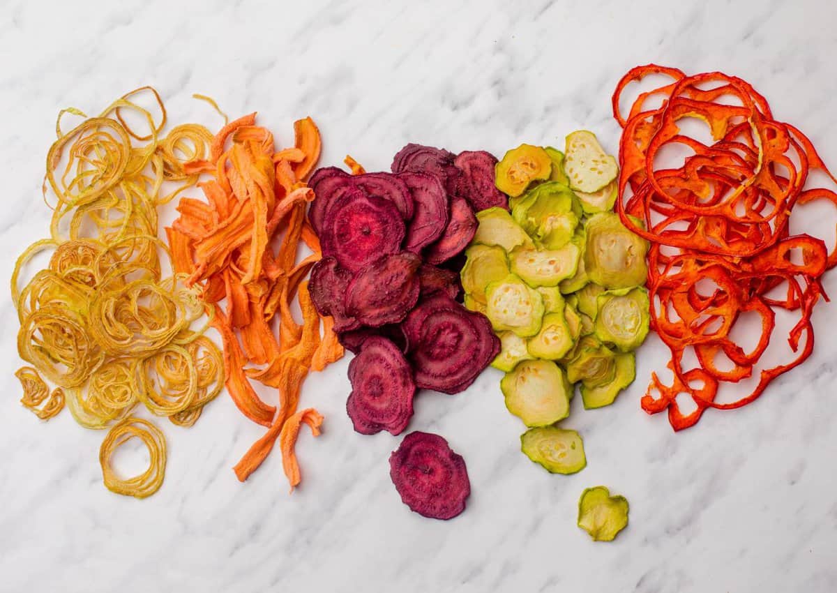An assortment of the best foods to dehydrate lies on a marble surface, featuring beet, carrot, zucchini, and bell pepper slices turned into crisp vegetable chips.