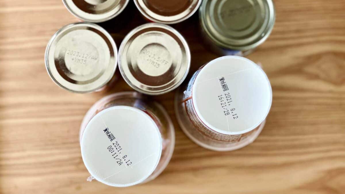 A group of canned food items arranged on a wooden surface, with visible expiration dates printed on the lids, ranging from 2023-12-24 to 2024-12-21.