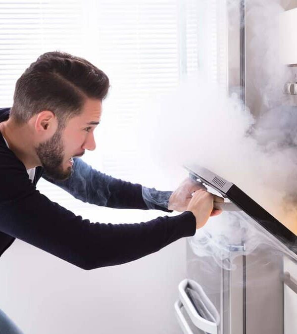 A man opens an oven door releasing smoke into the kitchen.