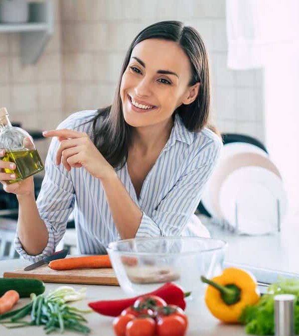 A woman in a kitchen holds a glass bottle of oil, smiling and pointing at it. Vegetables and kitchen tools are on the counter in front of her. A smartphone on a tripod records the scene.