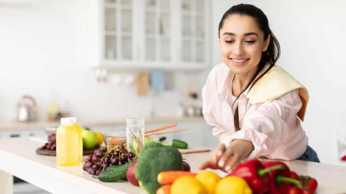 A woman in a kitchen reaches for produce on a counter filled with fruits and vegetables, including grapes, broccoli, bell peppers, and lemons.