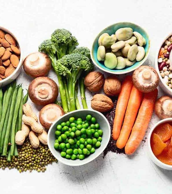 A variety of vegetables, nuts, seeds, and legumes are arranged on a white surface. Items include carrots, mushrooms, nuts, peas, beans, broccoli, and more.
