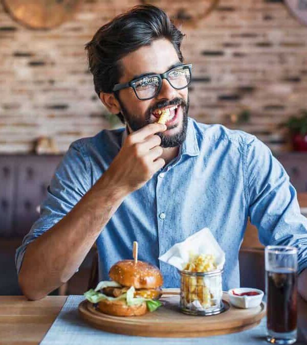 Man in glasses eating at a restaurant, with a burger, fries, glass of soda, and ketchup on the table.