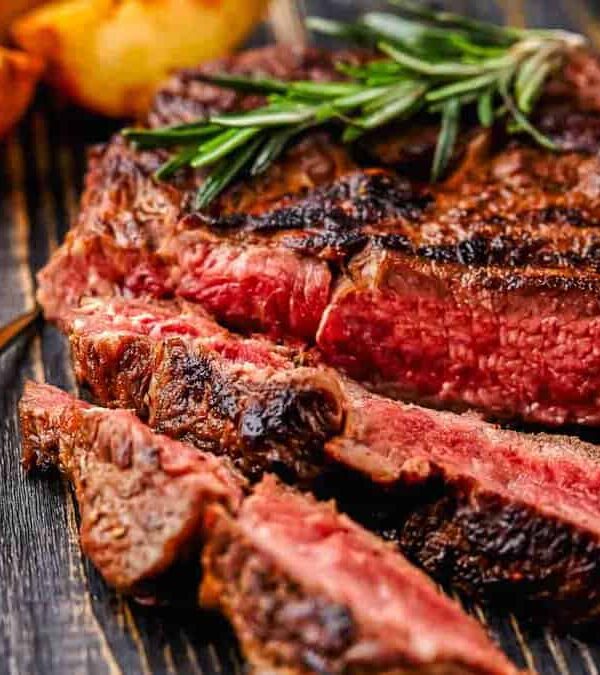 A sliced medium-rare steak garnished with rosemary on a wooden board, accompanied by roasted potatoes and a small bowl of sauce.