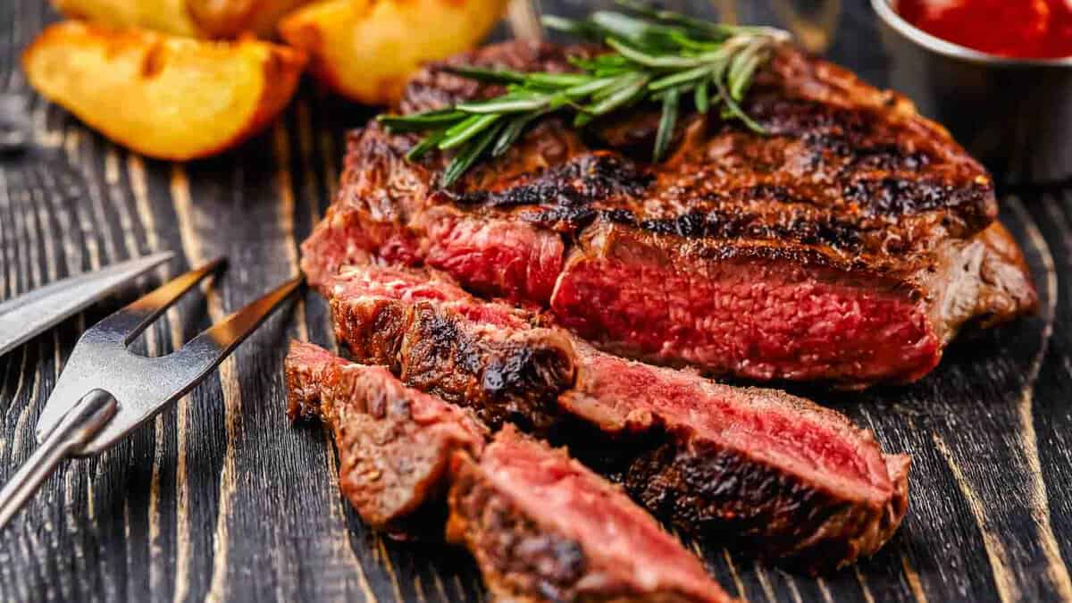 A sliced medium-rare steak garnished with rosemary on a wooden board, accompanied by roasted potatoes and a small bowl of sauce.