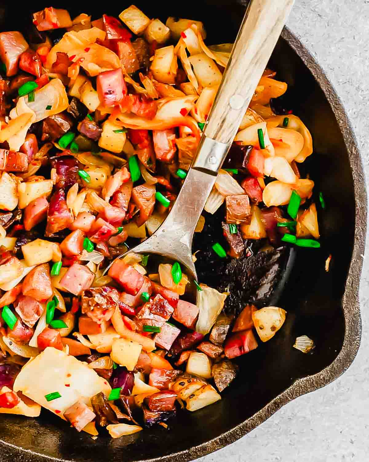 A skillet with a mixture of diced potatoes, onions, peppers, and ham, garnished with chopped green onions. A metal spatula is placed inside the skillet.