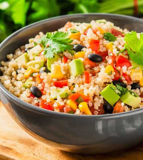 A gray bowl filled with quinoa, black beans, corn, avocado, tomatoes, and cilantro, placed on a wooden surface with a green checkered cloth and fresh vegetables in the background.