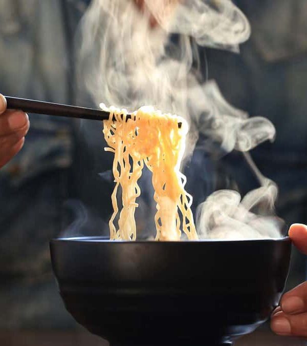 A person in denim shirt lifts steaming noodles from a black bowl using chopsticks.