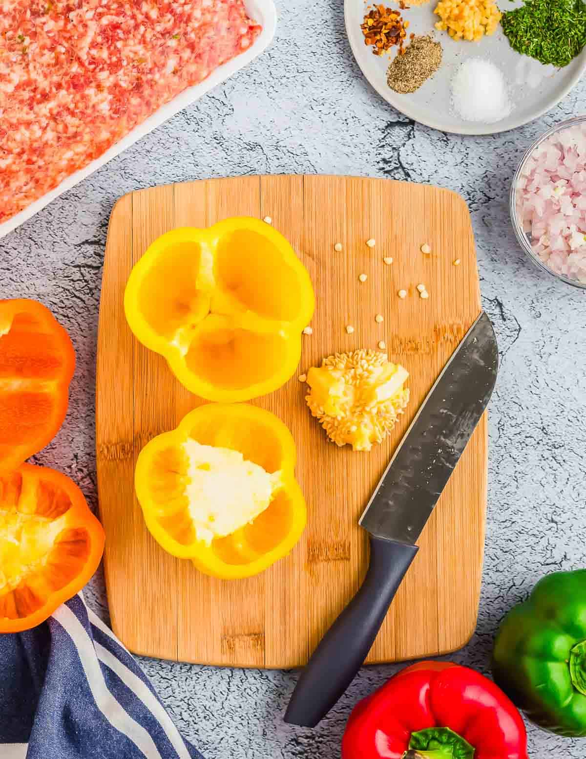 A yellow bell pepper cut in half with seeds removed sits on a wooden cutting board next to a knife. Ground sausage, seasonings, chopped onions, and other bell peppers are visible around the board.