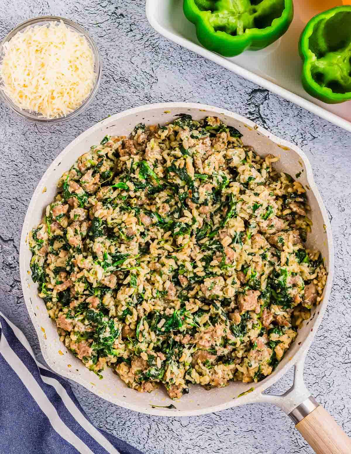 A skillet filled with a mixture of ground sausage, spinach, and rice, next to a bowl of shredded cheese and halved green bell peppers on a tray. A blue striped cloth is partially visible.