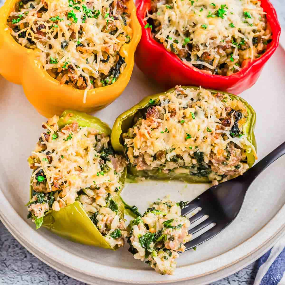 A plate with three Italian sausage stuffed bell peppers in yellow, red, and green. They are topped with melted cheese and herbs. A fork holds a bite taken from the green pepper.