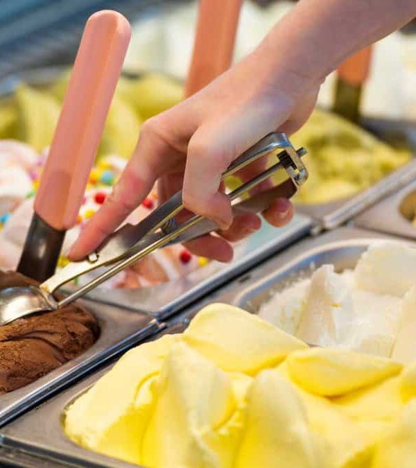 A hand scoops chocolate ice cream from a display case containing multiple flavors of ice cream or gelato.