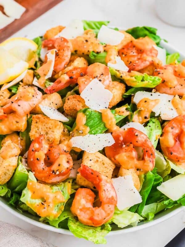 A fresh shrimp Caesar salad with romaine lettuce, shrimp, croutons, shaved parmesan cheese, and Caesar dressing is served in a white bowl with black utensils on a marble table.