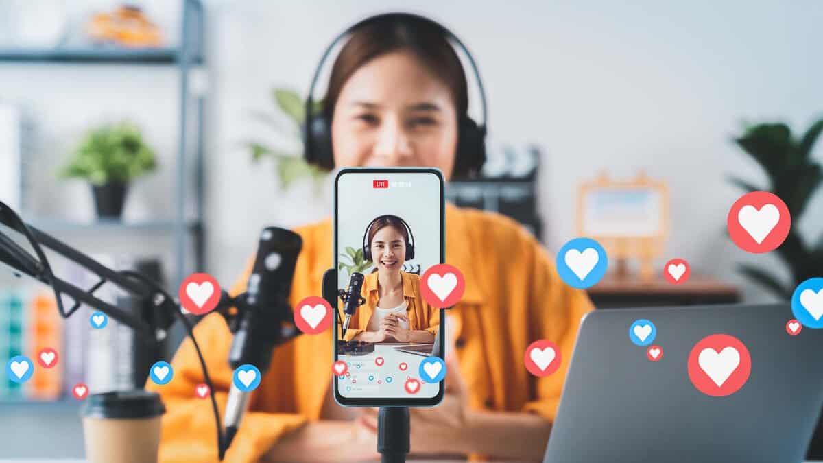 A person wearing headphones is live streaming with a microphone, viewed on a smartphone screen surrounded by floating heart and like icons. A laptop is on the table.