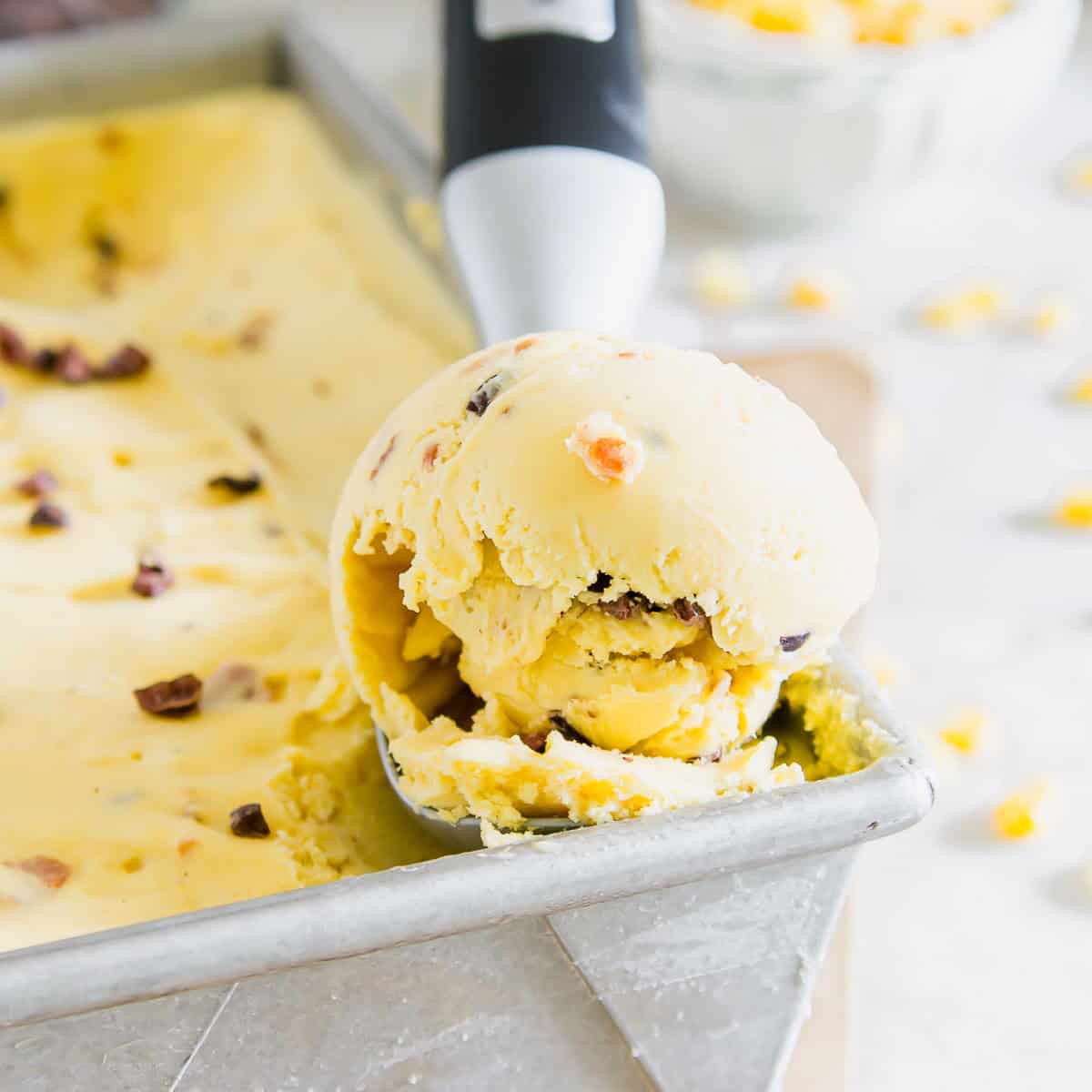 A metal ice cream scooper holds a scoop of sweet corn ice cream with small chocolate chips, over a rectangular metal tray filled with the same ice cream.
