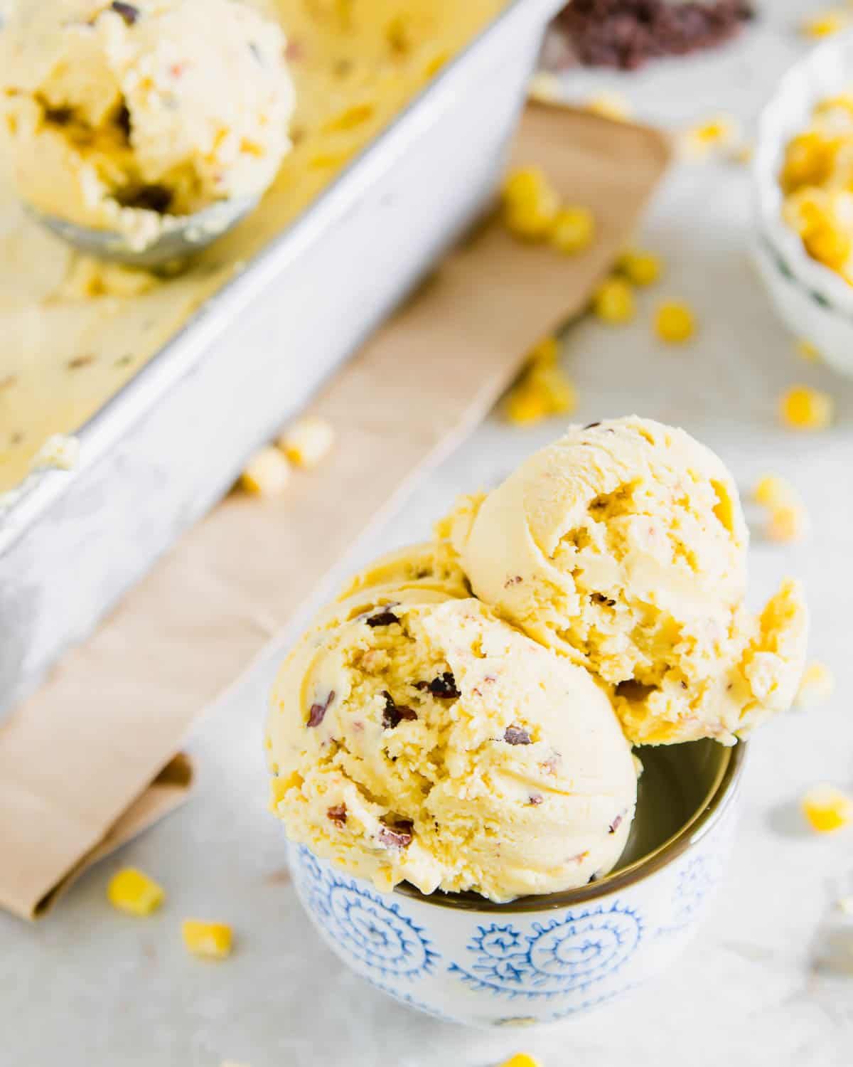 Two scoops of ice cream with corn kernels and chocolate chunks in a blue floral-patterned bowl, set on a light surface. A metal tray with more ice cream and scattered corn kernels is in the background.