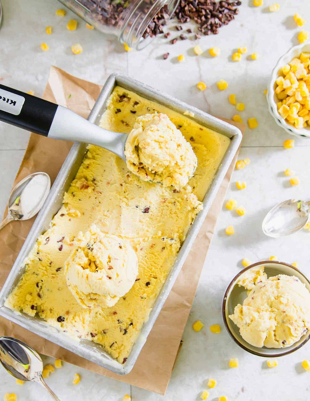 A rectangular container of corn ice cream with two scoops removed, surrounded by scattered corn kernels, spoons, and a bowl of ice cream on a light surface.