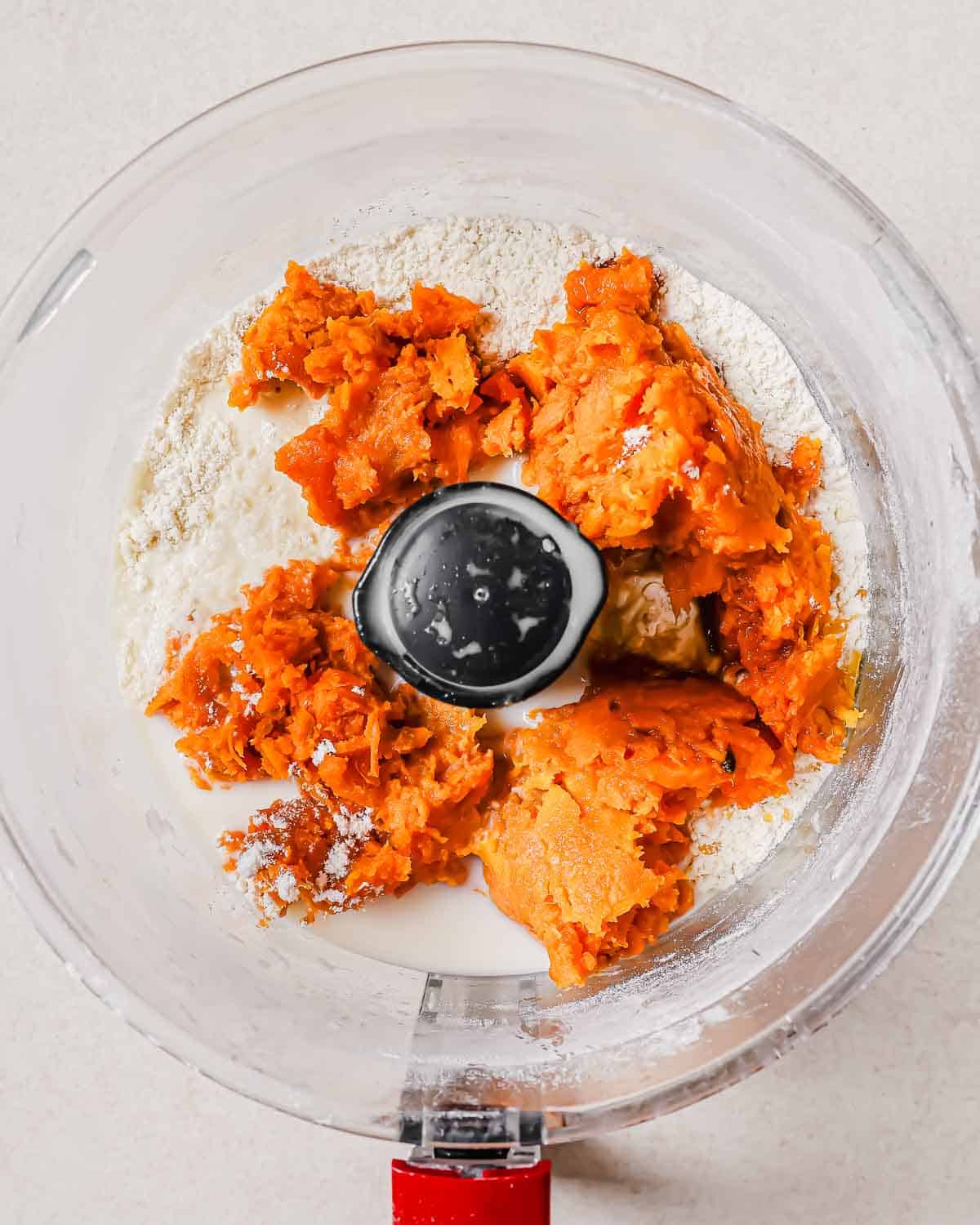 Top-down view of an open food processor containing flour and chunks of orange sweet potato.