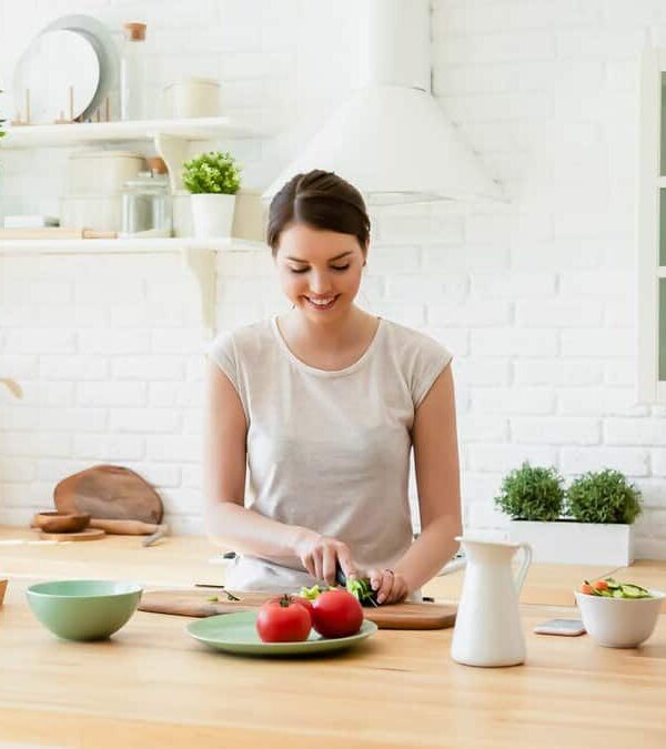 A person is standing at a wooden kitchen counter, chopping vegetables. The kitchen is bright with white cabinets and green plants.
