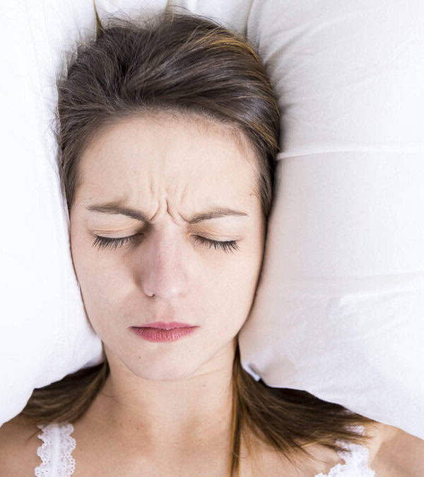 A woman lies in bed with a slightly pained expression, eyes closed, and her head between two white pillows, covering her ears.