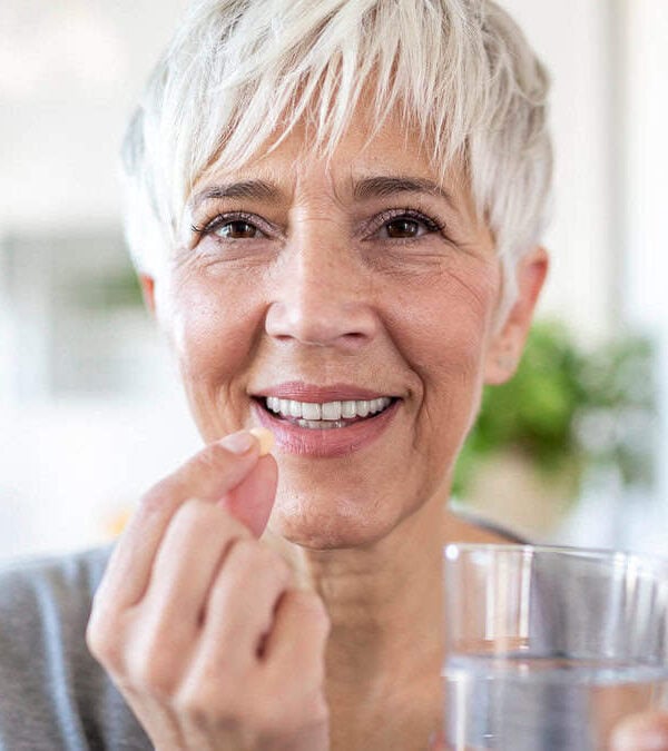 An elderly woman with short white hair smiles while holding a pill in one hand and a glass of water in the other.